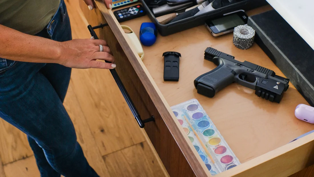 Mom looking into a drawer with remotes, household items, a watercolor paint set, and a pistol equipped with the B.O.S. Lock biometric firearm lock